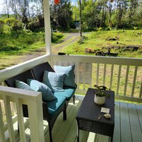 Un porche con un sofá de mimbre y una mesa, rodeado por una barandilla, con vista a un jardín con un sendero y vegetación.