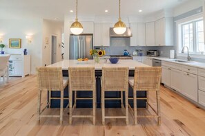 A modern kitchen with a central island, four wicker chairs, stainless steel appliances, and pendant lighting.