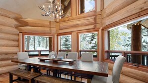 A log cabin interior with a large wooden dining table, multiple chairs, and large windows offering views of snow-covered trees. - Grand Alpine Lodge, Ski Access, Hot Tub, Sleeps 20 (Whitefish)