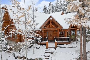 A wooden cabin with a snow-covered roof, surrounded by trees and a clear sky.