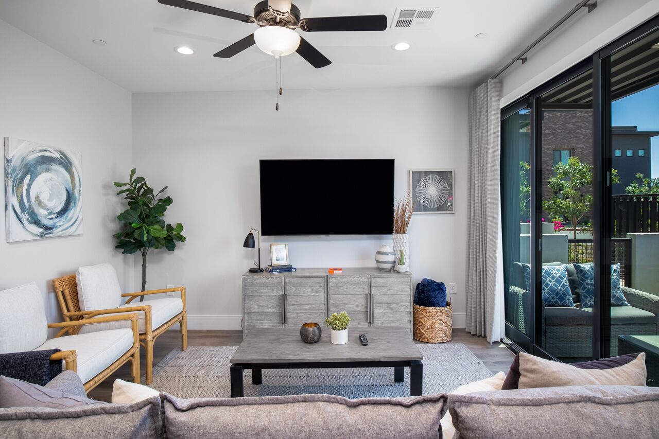 A modern living room with a flat-screen TV, a ceiling fan, and a large window offering a view of the outdoors.