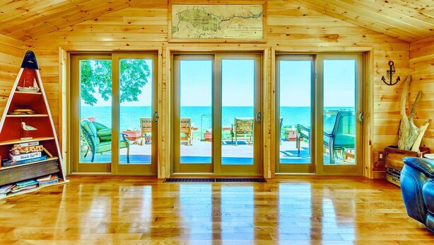 Una cabaña de madera con un amplio ventanal corredera que da a una terraza con mobiliario exterior y vistas al océano.