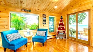 Interior de una cabaña de madera con dos sillones azules, una mesita y una escalera que lleva a un balcón con vista al océano.