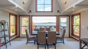 A dining room with a view of a lake, featuring a table with chairs, large windows, and a ceiling with exposed beams. - Luxury Lakefront Retreat, Hot Tub, Game Room, Views (Nisswa)