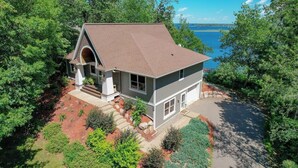 A two-story house with a sloped roof, a front porch, and a view of a lake. - Luxury Lakefront Retreat, Hot Tub, Game Room, Views (Nisswa)