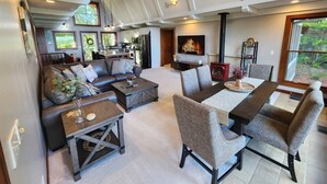 A spacious living room with a sofa, coffee table, and chairs, a dining area with a table and chairs, a kitchen in the background, and a large window with a view of greenery. - Luxury Lakefront Retreat, Hot Tub, Game Room, Views (Nisswa)