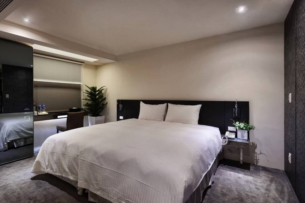 A modern hotel room with a large bed, a bedside table with a lamp, a mirror, and a potted plant.