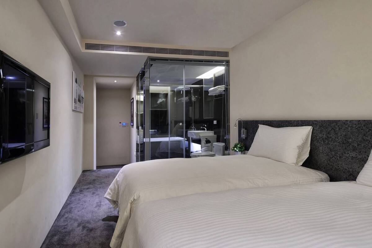 A hotel room with a large bed, a TV mounted on the wall, a bathroom with a glass shower enclosure, and a small bedside table.
