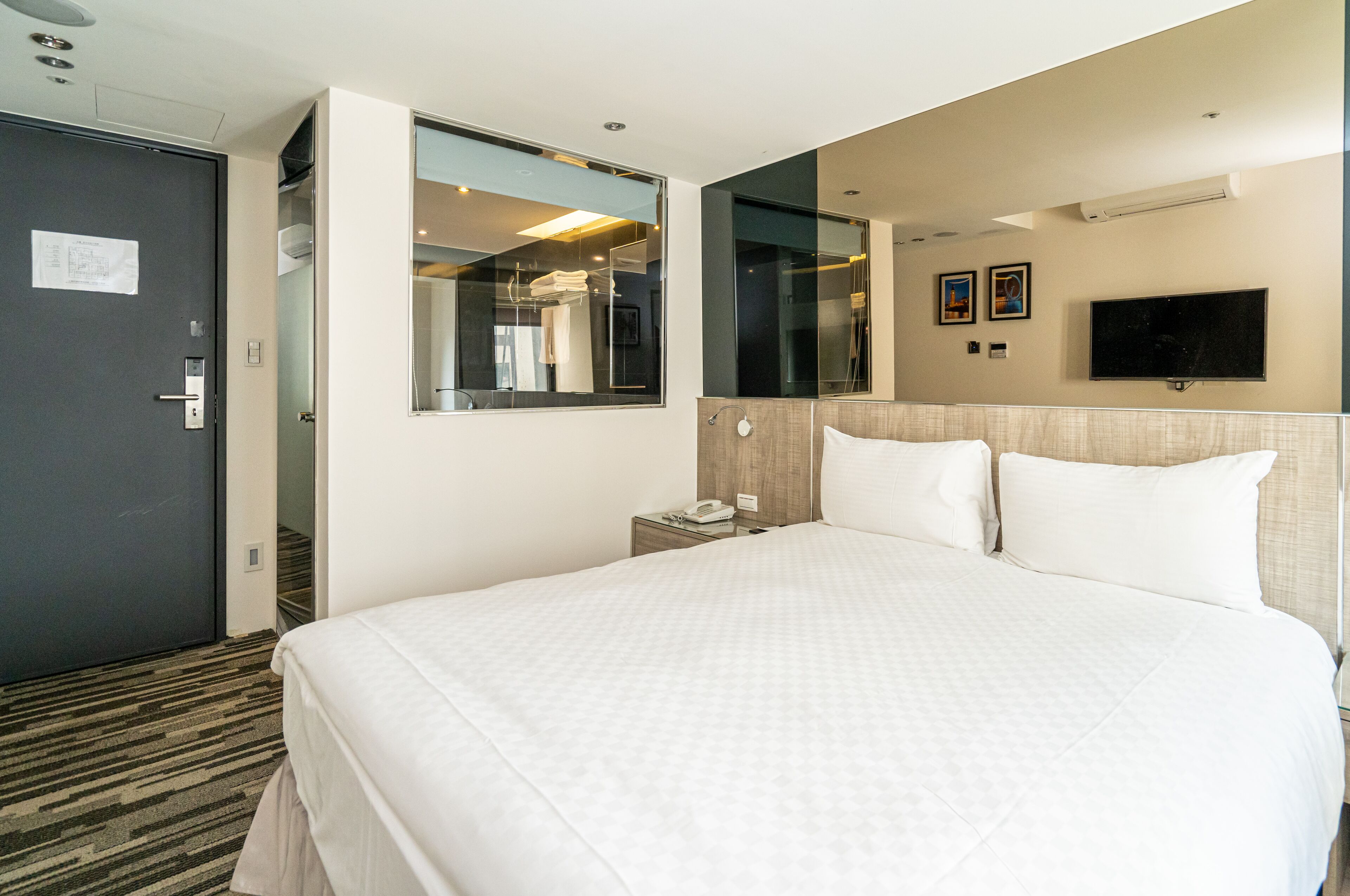 A hotel room with a large bed, a bedside table with a phone, a mirror, and a TV mounted on the wall.