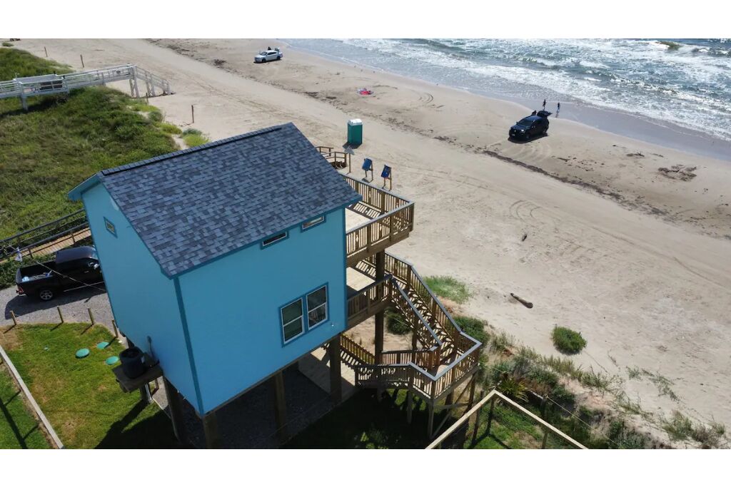 Un bâtiment bleu doté d’une terrasse et d’un escalier, situé près d’une plage où se trouvent quelques voitures et un poste de surveillance des sauveteurs.