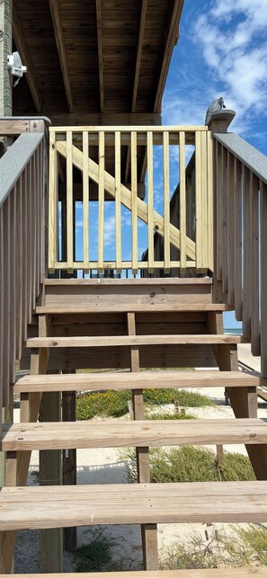 Un escalier en bois mène à une terrasse dotée d’une rambarde, qui offre une vue sur une plage de sable bordée de végétation.