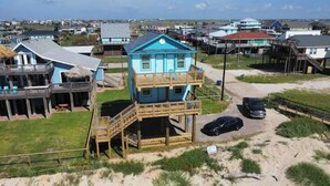 A two-story beach house with a balcony, a staircase, and a car parked nearby.