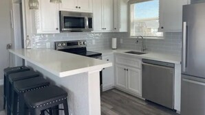 A modern kitchen with white cabinetry, a central island with stools, stainless steel appliances, and a window with blinds.