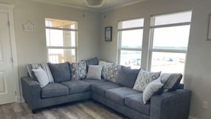 A grey sectional sofa with patterned cushions, a herringbone wood floor, and large windows with blinds.