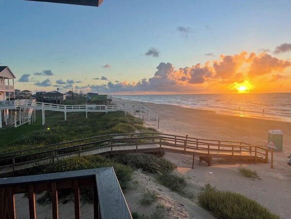 Vista sulla spiaggia con una passerella in legno che conduce alla spiaggia, case sullo sfondo e un tramonto sull'oceano.