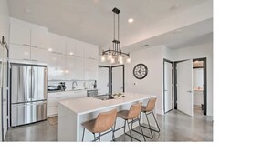 A modern kitchen with white cabinetry, a stainless steel refrigerator, a central island with bar stools, and a pendant light.