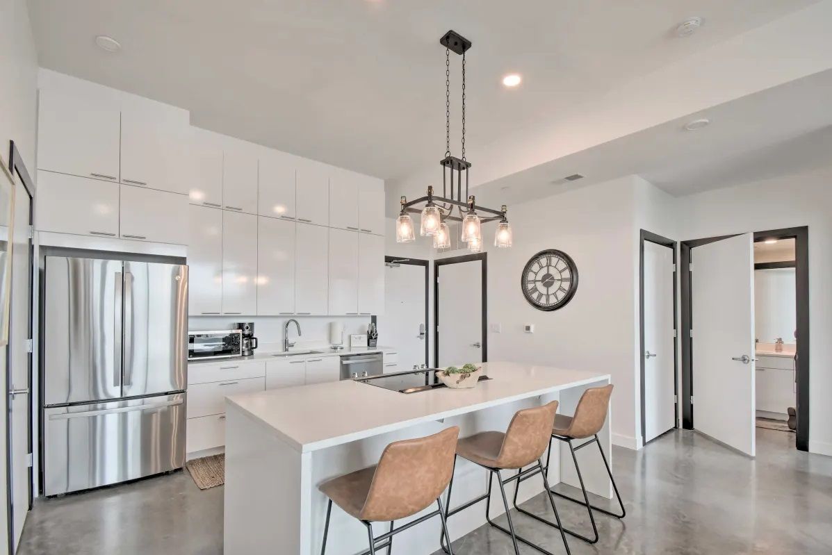 A modern kitchen with white cabinetry, a stainless steel refrigerator, a central island with bar stools, and a pendant light.