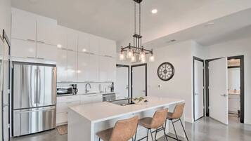 A modern kitchen with white cabinetry, a stainless steel refrigerator, a central island with bar stools, and a pendant light.