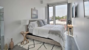A modern bedroom with a large bed, a side table, a floor lamp, and a view of the city through large windows.