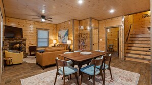 A cozy cabin interior with a wooden dining table, chairs, a sofa, a fireplace, and a staircase.