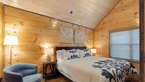 A wooden cabin bedroom with a bed, two wall-mounted lamps, a chair, and a window with blinds.