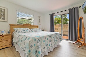 A bedroom with a bed, a wooden dresser, a bedside table, a mirror, and a view through a sliding glass door.