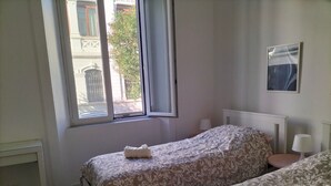 Ein Schlafzimmer mit zwei Betten, einem Fenster, einem Nachttisch, einer Lampe und einem gerahmten Bild an der Wand.