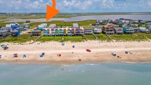 Ein Luftbild einer Küstenstadt mit Häusern auf Stelzen über dem Wasser, einem Sandstrand mit Sonnenschirmen und einem klaren Himmel.
