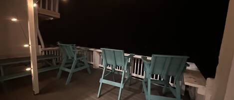 A covered outdoor seating area with wooden chairs and tables, string lights, and a railing.