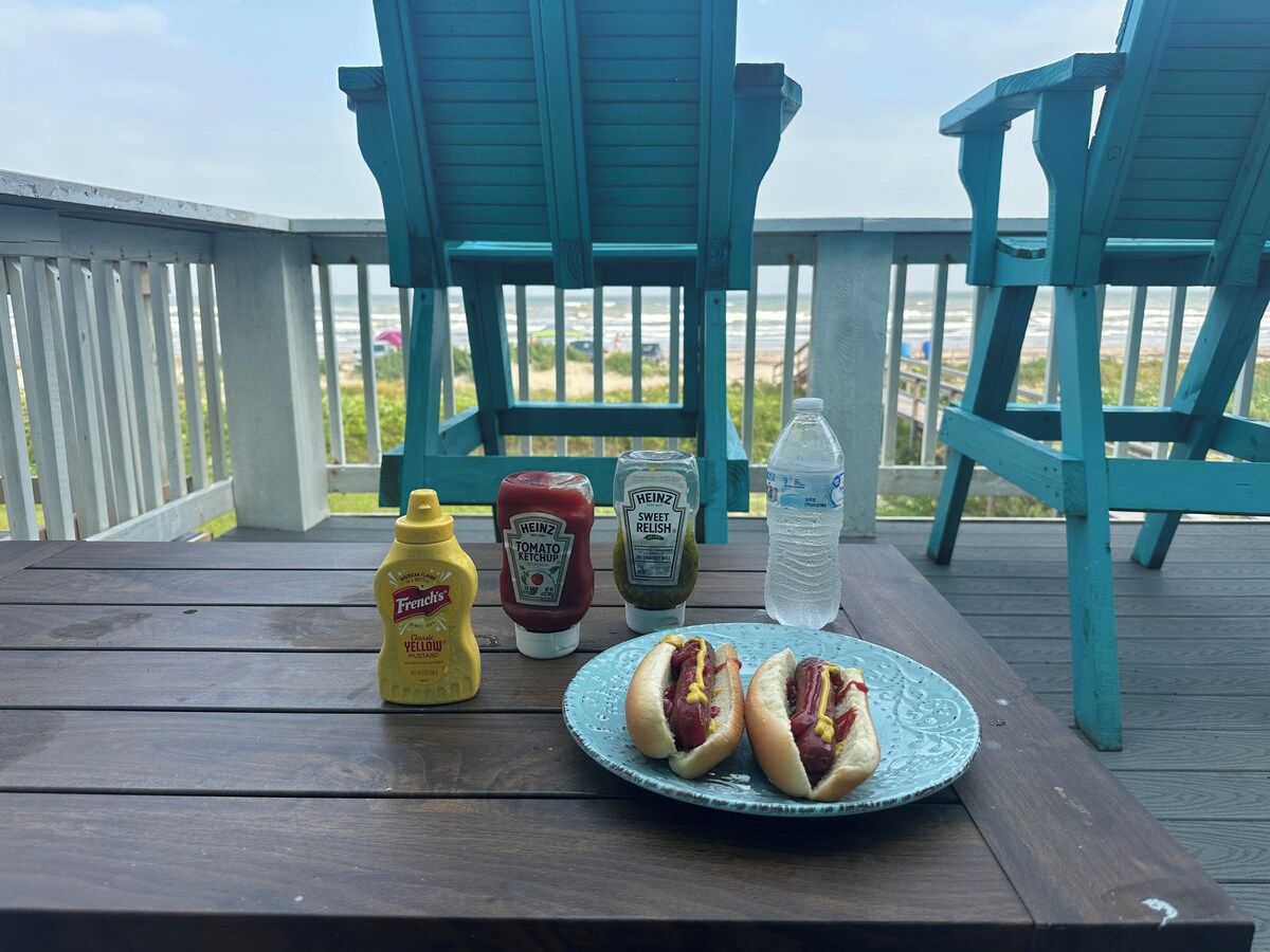 Een houten terras met twee blauwe Adirondack-stoelen, een picknicktafel en een bord met twee hotdogs.