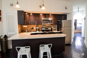 A modern kitchen with dark cabinets, a central island, two bar stools, a microwave, a stove, and a refrigerator.