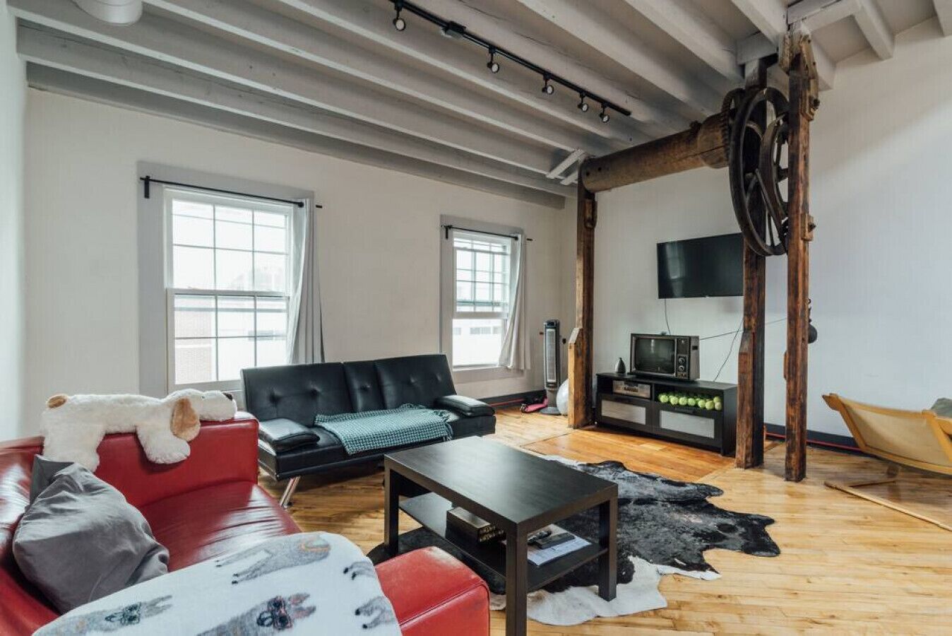 Een ruime woonkamer met een rode bank, een zwarte leren hoekbank en een houten salontafel. Er is een televisie op een houten dressoir, een groot raam en een balkenplafond.
