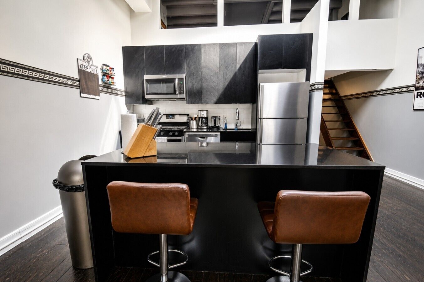A modern kitchen with a black island, stainless steel appliances, and two leather bar stools.
