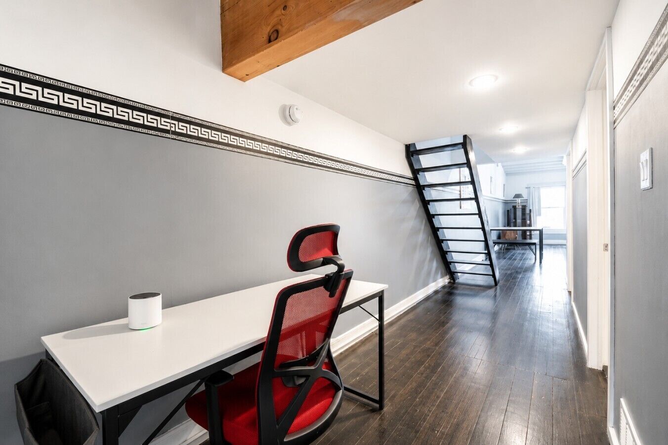 Un interior modern amb una taula blanca, una cadira vermella i una escala negra adossada a una paret blanca amb un detall decoratiu.