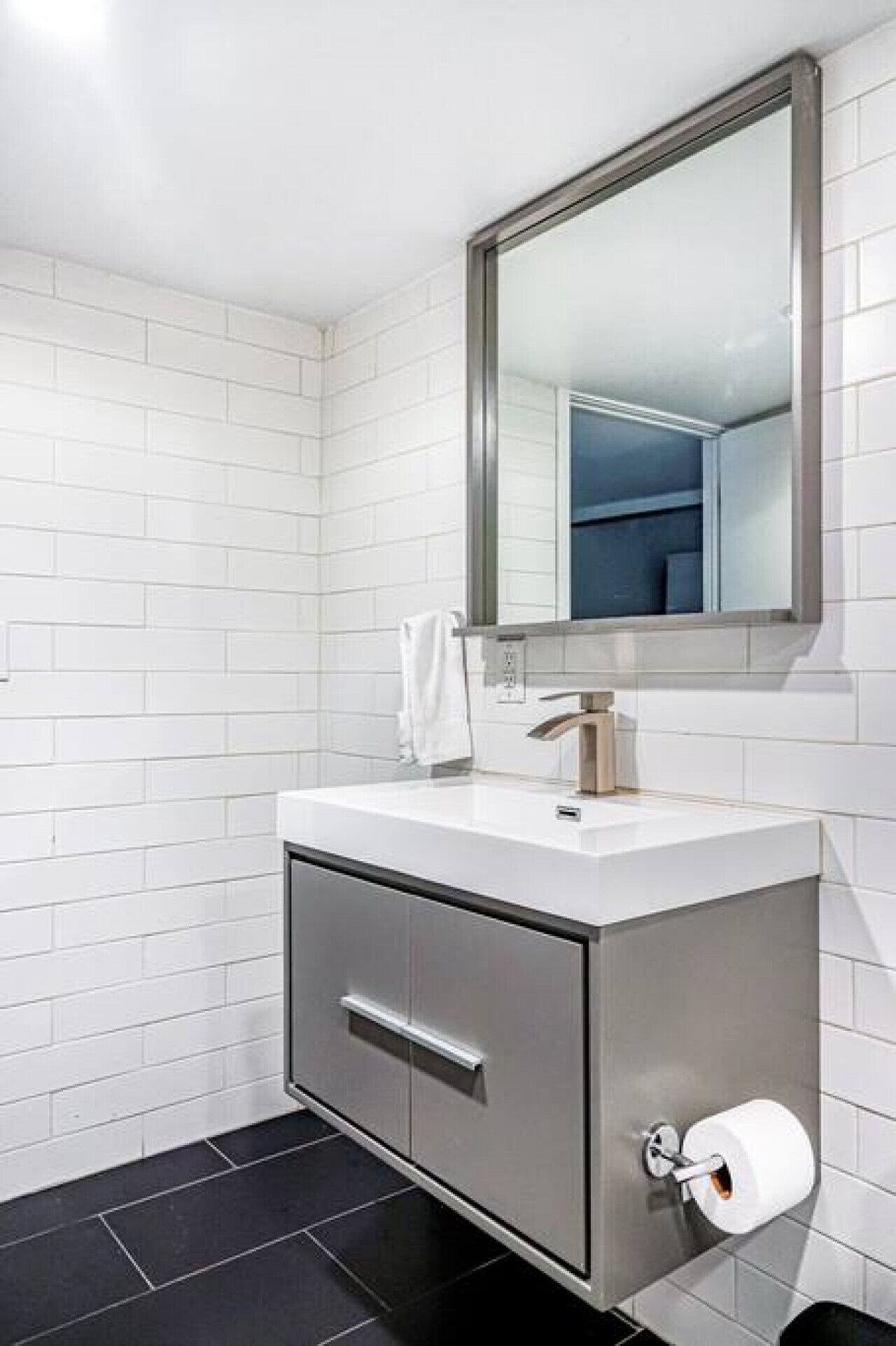 Un baño moderno con azulejos blancos tipo metro, un lavamanos blanco con grifo plateado, un espejo sobre el lavamanos y un inodoro con un dispensador de toallas de papel.