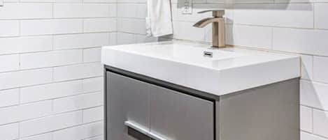 Un baño moderno con azulejos blancos tipo metro, un lavamanos blanco con grifo plateado, un espejo sobre el lavamanos y un inodoro con un dispensador de toallas de papel.