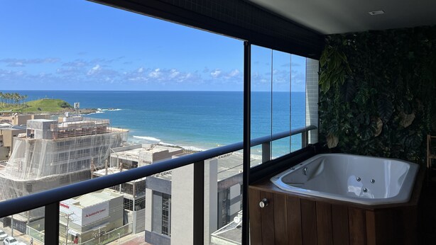A balcony with a view of the ocean and cityscape, a wooden bathtub, and a green wall. - Sofisticado Loft Vista Mar - Farol da Barra (Salvador)