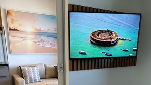 A hotel room with a bed, a TV mounted on the wall displaying a tropical scene, a sofa, and a view of the sea. - Sofisticado Loft Vista Mar - Farol da Barra (Salvador)