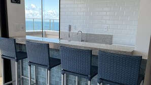 A modern kitchen with a white brick backsplash, a large window overlooking the ocean, and three bar stools with woven tops. - Sofisticado Loft Vista Mar - Farol da Barra (Salvador)