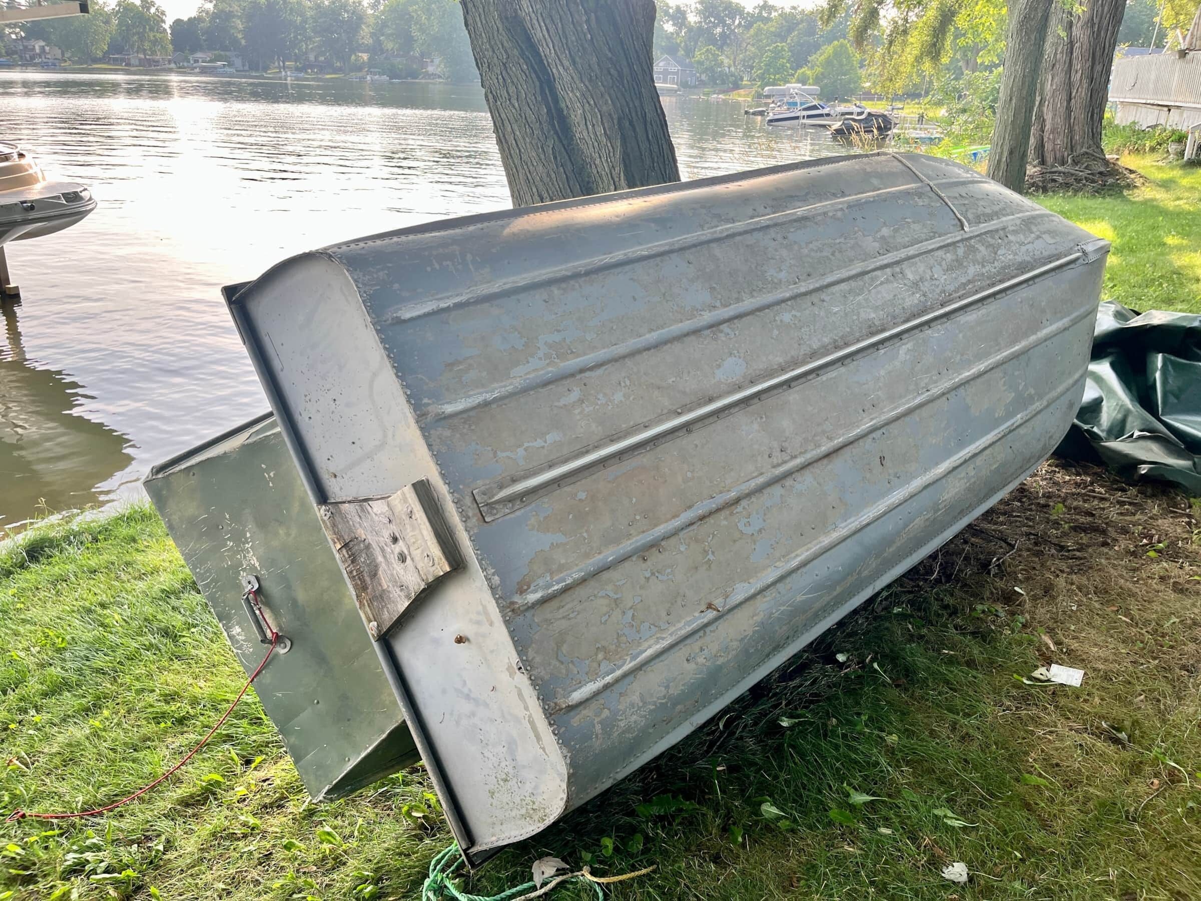 Une structure métallique, peut-être un bateau, reposant sur de l’herbe, près d’une étendue d’eau, avec des arbres et des maisons en arrière-plan.
