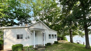 A white single-story house with a porch, a small deck, and a view of a lake. - Cottages on Sand Lake - The Lazy Lakehouse (Nottawa)