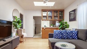 Un salon moderne avec un canapé gris, une petite table basse blanche et un téléviseur posé sur un meuble en bois. On aperçoit également un coin cuisine avec des meubles en bois et une table à manger à l’arrière-plan.