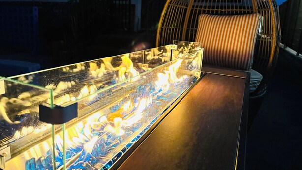 Une pièce avec une table en verre sur laquelle est disposé un élément décoratif représentant un foyer, une chaise en osier et un escalier en arrière-plan.