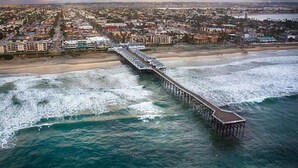 Un paysage urbain côtier avec une longue jetée qui s’avance dans l’océan, entouré d’immeubles résidentiels et d’une plage.