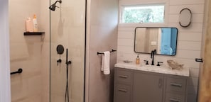 Un baño con ducha envuelta en vidrio, tocador con encimera de mármol, espejo y ventana sobre el lavabo.