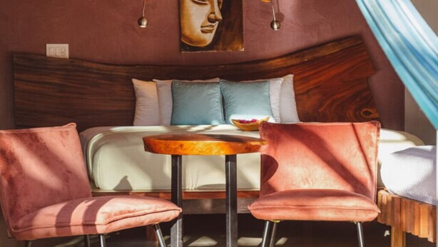 A bedroom with a wooden headboard, a bed with white and blue bedding, two pink chairs, a small round table, and a Buddha painting on the wall. - Beach Front Earth Suites #1 (Playa Guiones)
