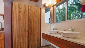 A bathroom with two sinks, wooden cabinets, a large mirror, and a window with a view of palm trees. - Beach Front Earth Suites #1 (Playa Guiones)