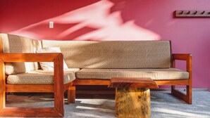 A wooden sofa with a patterned cushion against a pink wall, a wooden coffee table, and a wooden shelf with a light fixture. - Beach Front Earth Suites #1 (Playa Guiones)