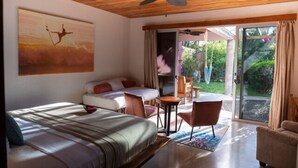 A bedroom with a bed, a desk, and a chair, a balcony with a view of the garden, and a ceiling fan. - Beach Front Earth Suites #1 (Playa Guiones)