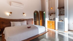 Quarto com cama grande, cabeceira de madeira e mesa de cabeceira pequena. Cozinha visível através de uma porta aberta.